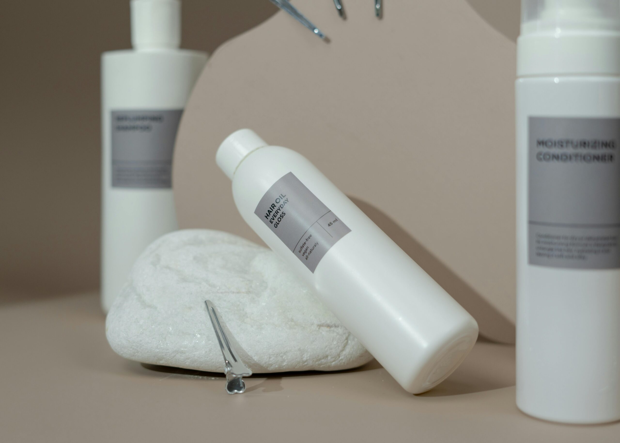 Collection of white cosmetic bottles with gray labels, including shampoo, conditioner, and hair oil, displayed on a beige surface with a decorative stone and hair clips.