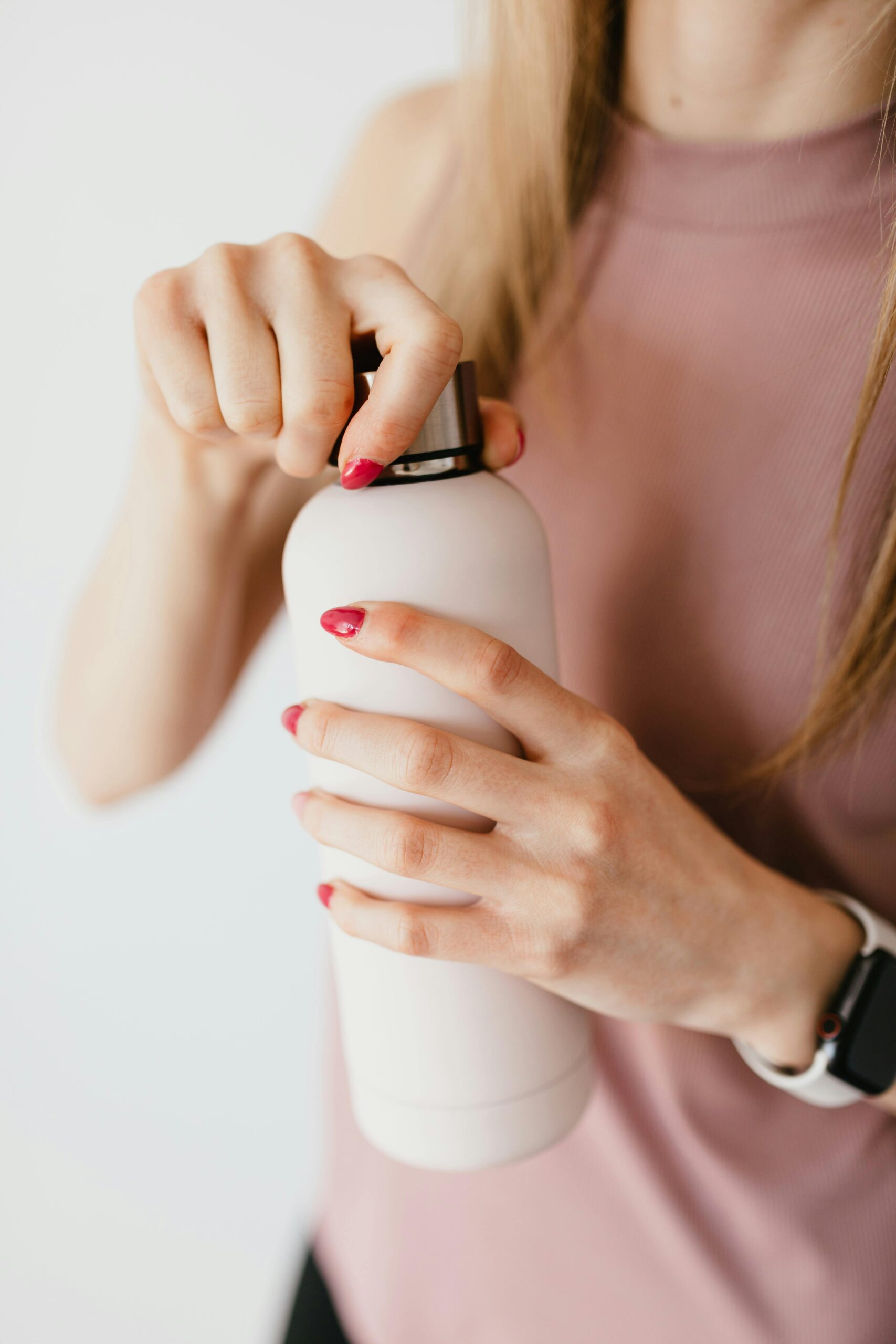Close-up of a woman’s hands with red nail polish holding a white pump bottle, wearing a sleeveless top and a smartwatch.
